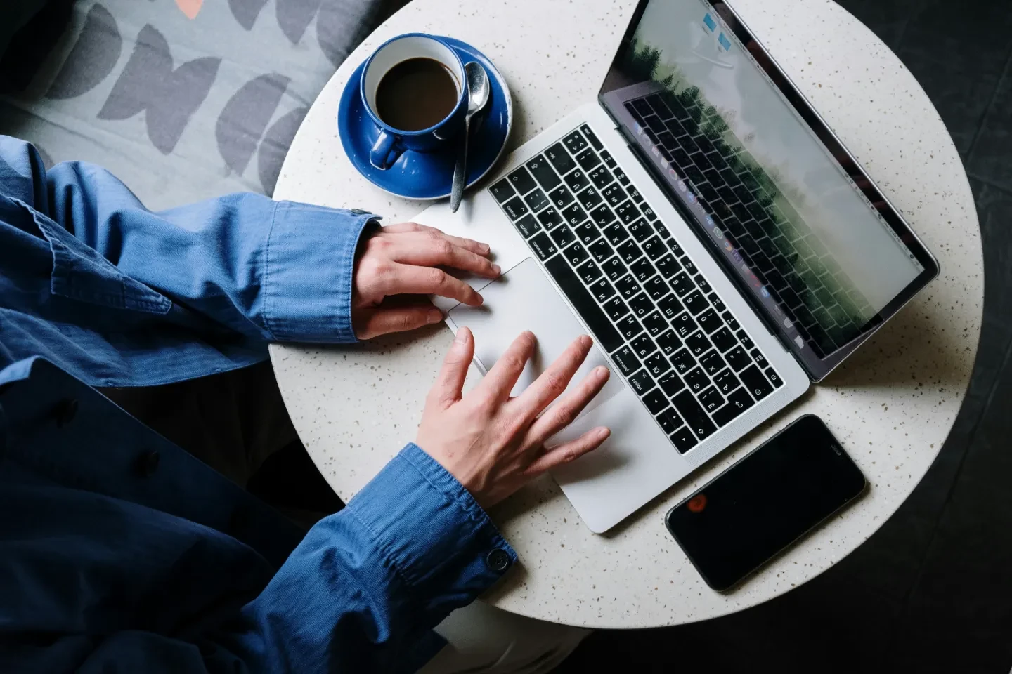 Two hands on a laptop touch pad with a cup of coffee on the left and a phone on the right side.