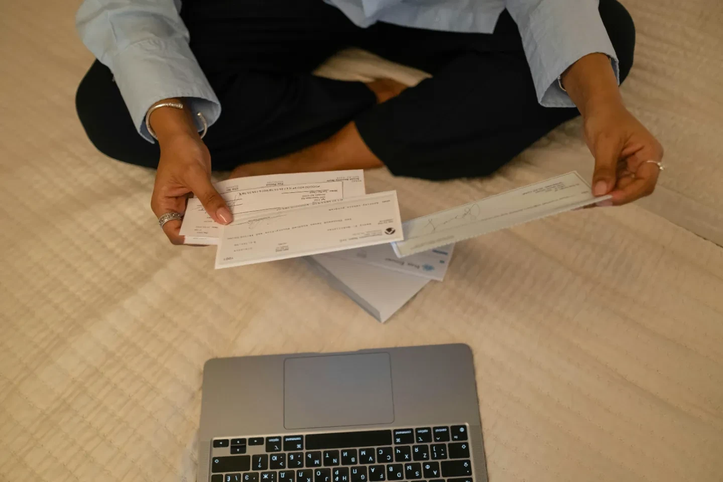 A person sitting cross-legged with checks in hand and laptop in front.