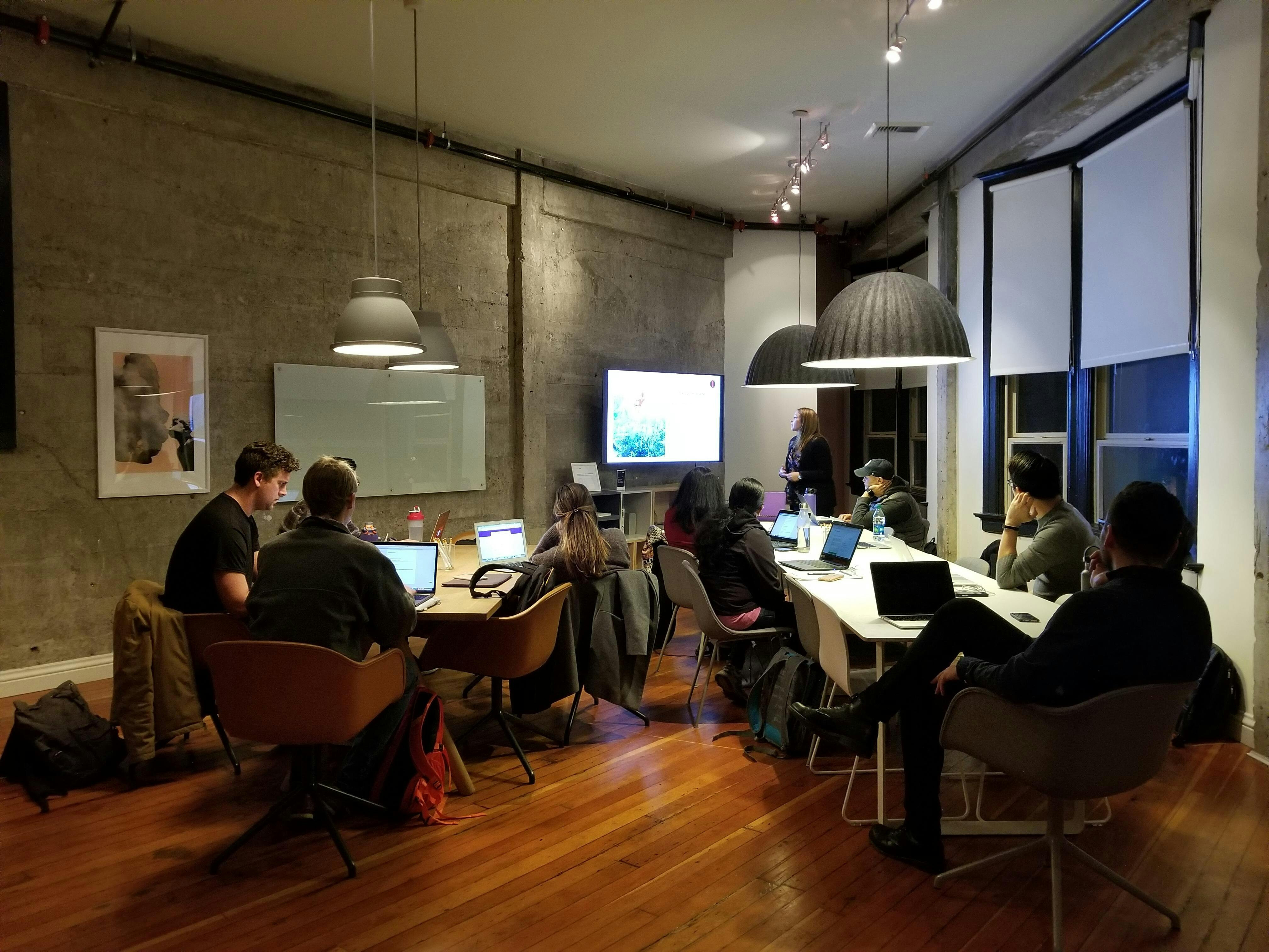 An office space with people sitting around two tables, looking at a presentation on a screen.